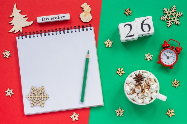 Winter composition. Wooden calendar Cup of cocoa with marshmallow, empty open notepad with pencil, snowflake, alarm clock on red and green background. Top view Flat lay Mockup