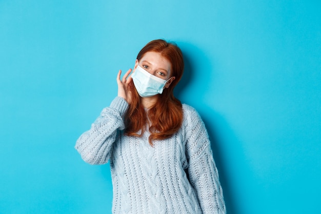 Winter, covid-19 and social distancing concept. Cute redhead teenage girl, wearing face mask and tuck hair strand behind ear, staring at camera, standing over blue background.