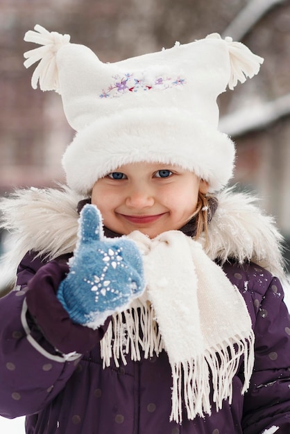 Winter face positive smiling winter portrait happy girl kid child in white hat shows thumb up like