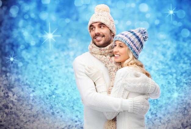 winter, fashion, couple, christmas and people concept - smiling man and woman in hats and scarf hugging over blue glitter or lights background