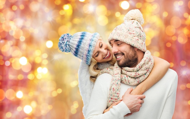 winter, fashion, couple, christmas and people concept - smiling man and woman in hats and scarf hugging over lights background