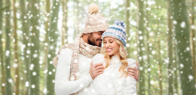 winter, fashion, couple, christmas and people concept - smiling man and woman in hats and scarf hugging over snowflakes and forest background