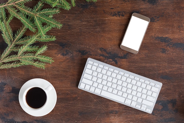 Winter green fir tree branches with cup of tea, mobile phone and white computer keyboard on wooden backgrond, flat lay and view from above