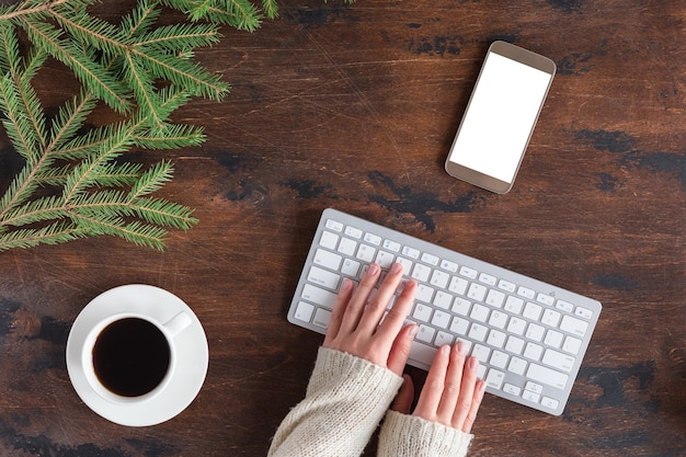 Winter green fir tree branches with cup of tea, mobile phone and white computer keyboard on wooden backgrond, flat lay and view from above