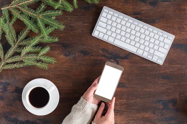 Winter green fir tree branches with cup of tea, mobile phone and white computer keyboard on wooden backgrond, flat lay and view from above