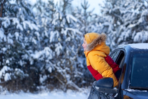 Winter holiday Boy child is leaning out of the car window and enjoying snow road and forest winter