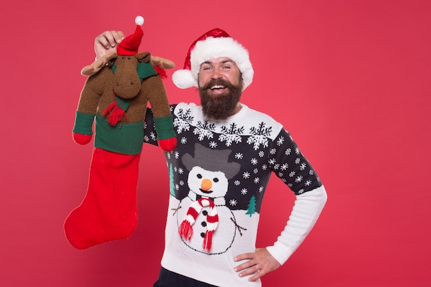 Winter holiday. Check contents of christmas stocking. Man in santa hat hold christmas gift red background. Hipster cheerful face got gift in christmas sock. Christmas stocking concept. Reindeer toy.