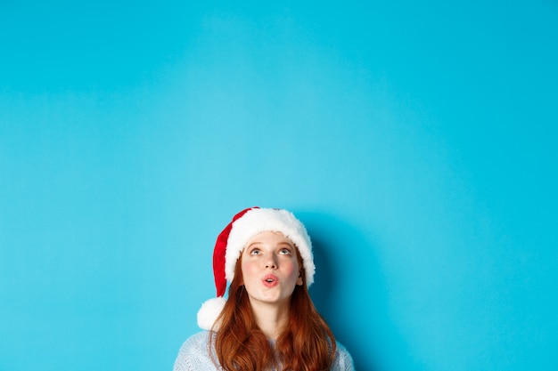 Winter holidays and Christmas eve concept. Head of pretty redhead girl in santa hat, appear from bottom and looking up at logo impressed, seeing promo offer, blue background