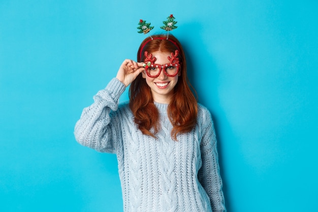 Winter holidays and Christmas sales concept. Beautiful redhead female model celebrating New Year, wearing funny party headband and glasses, smiling at camera.