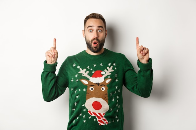 Winter holidays and christmas. Surprised man pointing fingers up, showing New Year promo offer, standing over white background