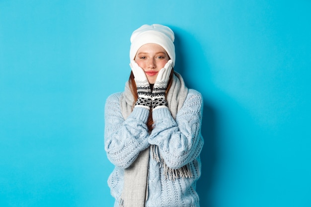 Winter and holidays concept. Cute girl in white beanie, sweater and gloves, squeezing cheeks and smiling pleased, warm-up after cold outdoors, standing over blue background.