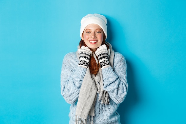 Winter and holidays concept. Cute redhead girl in white beanie and gloves smiling at camera, looking delighted, standing against blue background.