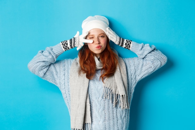 Winter and holidays concept. Happy redhead girl in beanie hat, scarf and gloves showing peace sign and pucker lips sassy, staring at camera, wearing sweater, posing over blue background.