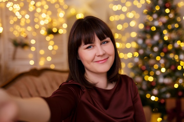 Winter holidays concept - young beautiful woman taking selfie photo in decorated living room with Christmas tree and festive led lights