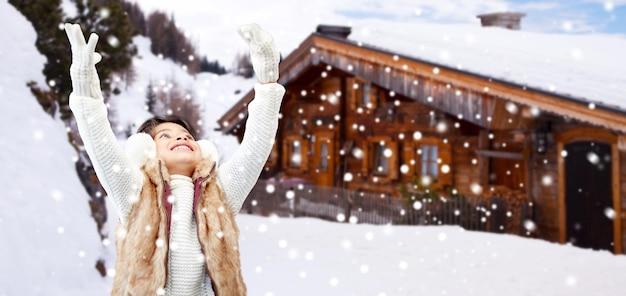 winter holidays, season, christmas, people and children concept - happy little girl wearing earmuffs and gloves over wooden country house background and snow