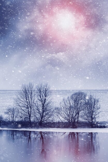 Winter landscape with trees by the river during a snowfall in the evening during sunset. Reflection of trees in the river in winter