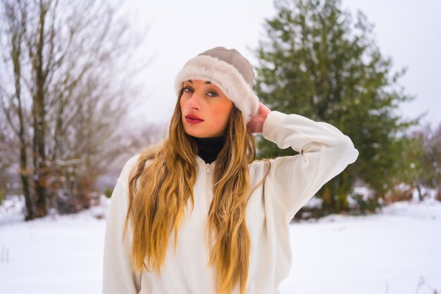 Winter lifestyle, portrait caucasian blonde with white winter outfit and woolen hat enjoying the snow next to snowy pine trees, vacation in nature