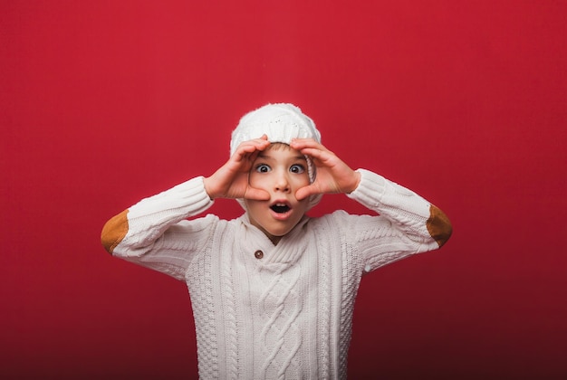 Winter portrait of a happy joyful childa boy in a knitted hat and sweater having fun on a red background the boy looks at his palms