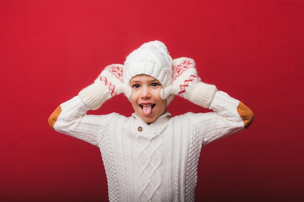 Winter portrait of a happy joyful childa boy in a knitted hat and sweater having fun on a red background the boy looks at his palms