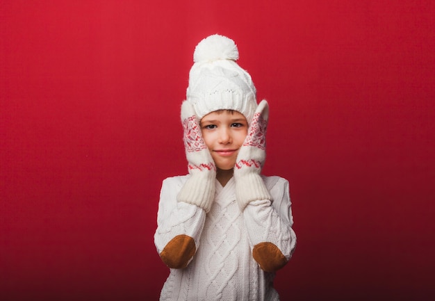 Winter portrait of a happy joyful childa boy in a knitted hat and sweater having fun on a red background the boy looks at his palms