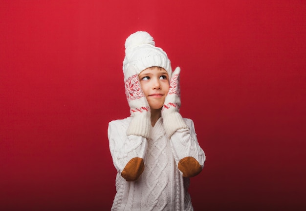 Winter portrait of a happy joyful childa boy in a knitted hat and sweater having fun on a red background the boy looks at his palms