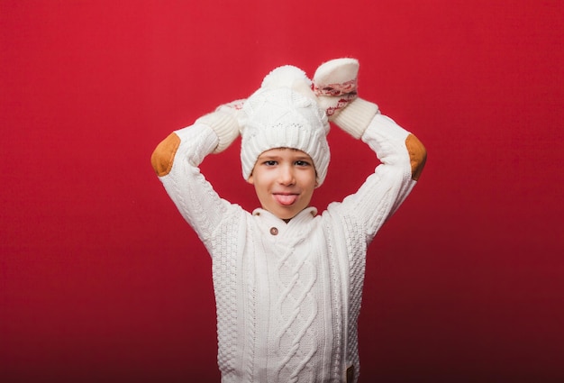 Winter portrait of a happy joyful childa boy in a knitted hat and sweater having fun on a red background the boy looks at his palms