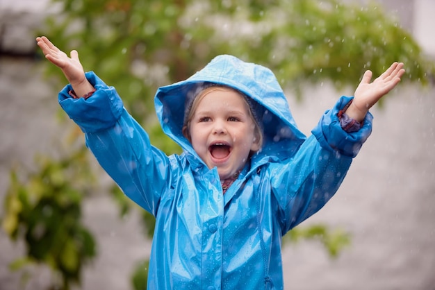 Photo winter raincoat and a happy girl playing in the rain outdoor alone having fun during the cold season kids water or wet with an adorable little female child standing arms outstretched outside