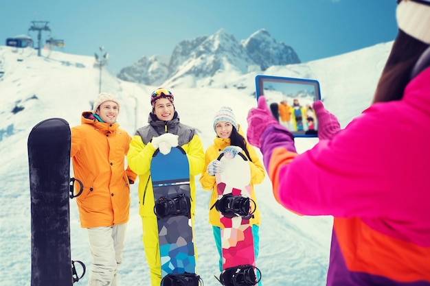 winter sport, technology, leisure, friendship and people concept - happy friends with snowboards and tablet pc computer taking picture over snow and mountain background