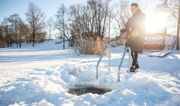 Winter swimming preparation Man standing by freshly cut ice hole on a snowy lakeside