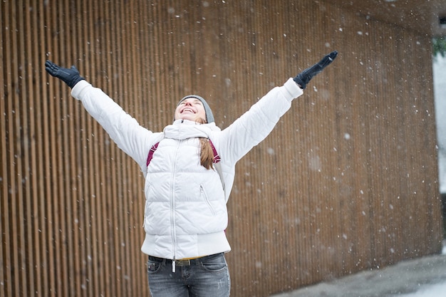 Winter time. Young happy caucasian girl wearing white jacket enjoying first snow with rised hands.