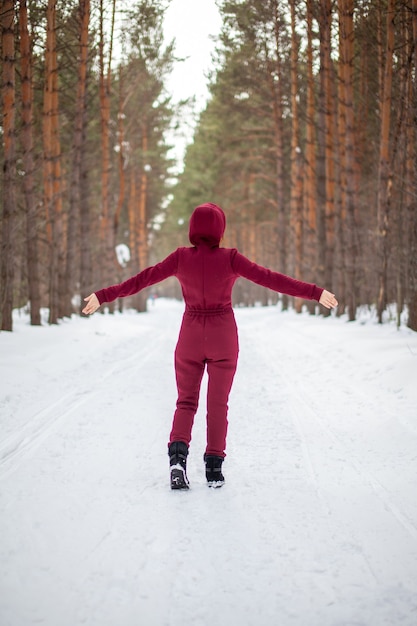 Winter walk in a snow-covered forest, A girl with a red jumpsuit and jacket walks among tall trees in nature.