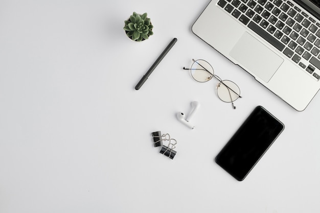 Wireless earphones, eyeglasses, pen, clips, smartphone, small domestic plant and laptop keypad on white desk