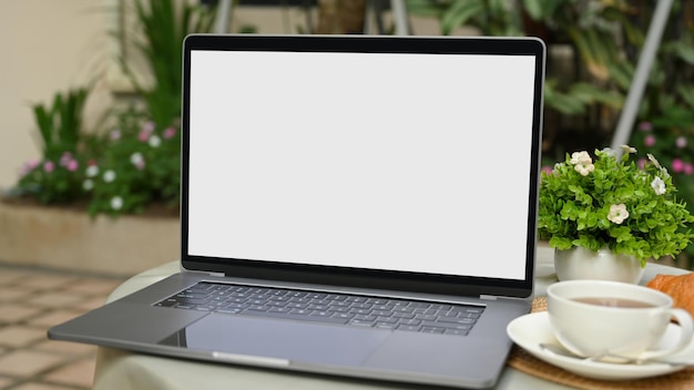 A wireless portable notebook laptop computer white screen mockup and coffee cup on coffee table in the outdoor cafe.