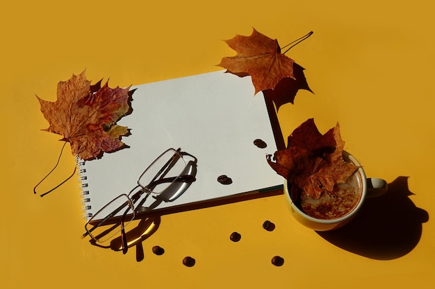 Wishes for a cozy autumn notebook and eyeglasses scattered maple leaves a cup of coffee with a maple leaf in it coffee beans on a yellow background top view