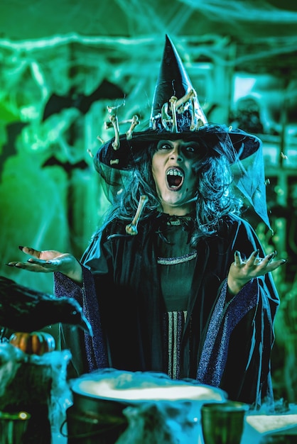 Witch with awfully face in creepy surroundings and smoky green background talks magic words to bones around her hat, above boiling cauldron. Halloween concept.