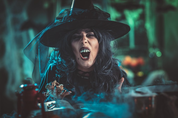 Witch with awfully face in creepy surroundings and smoky green background talks magic words to bones above boiling cauldron. Halloween concept.