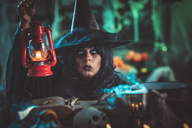 Witch with awfully face in creepy surroundings and smoky green background talks magic words to bones above boiling cauldron. Halloween concept.