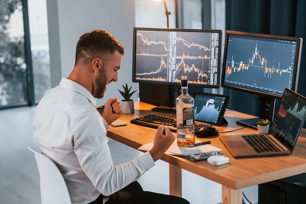 With alcohol Young businessman in formal clothes is in office with multiple screens