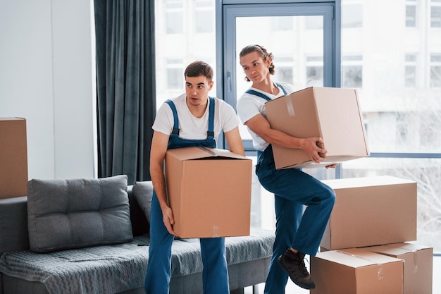 With boxes in hands Two young movers in blue uniform working indoors in the room