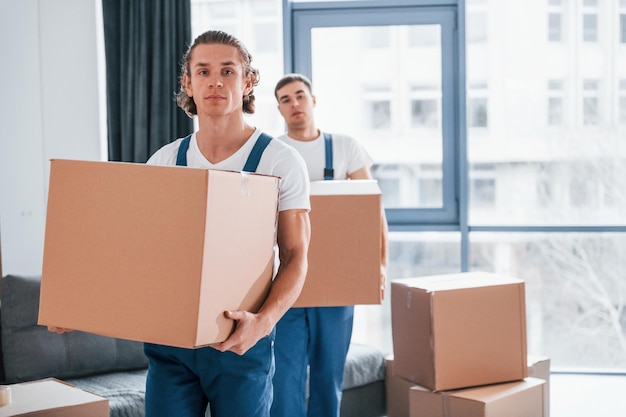 With boxes in hands Two young movers in blue uniform working indoors in the room