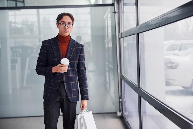 With cup of drink and shopping bags. Young business man in luxury suit and formal clothes is indoors in the office.