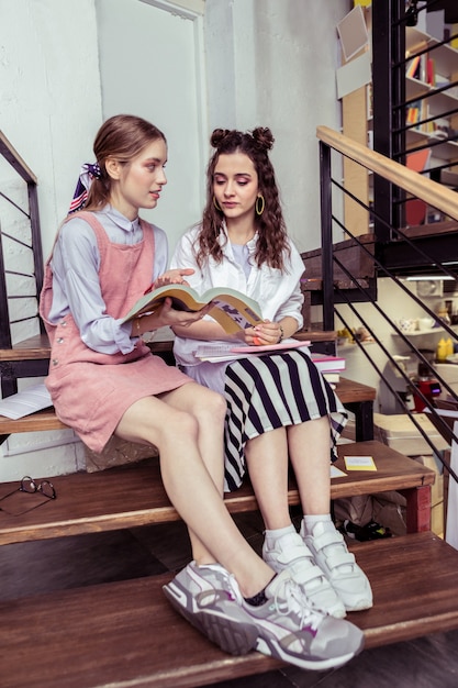 With curiousness. Fashionable tidy girls wearing sneakers while sitting on wooden stairs and discussing information in book