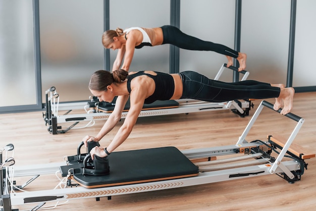 With help of gym equipment Two women in sportive wear and with slim bodies have fitness yoga day indoors together