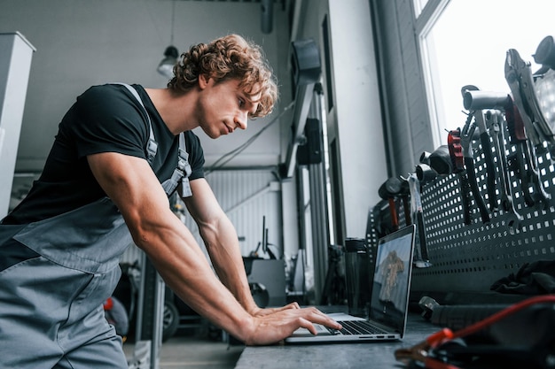 Photo with laptop adult man in grey colored uniform works in the automobile salon