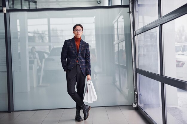 With shopping bags. Young business man in luxury suit and formal clothes is indoors in the office.