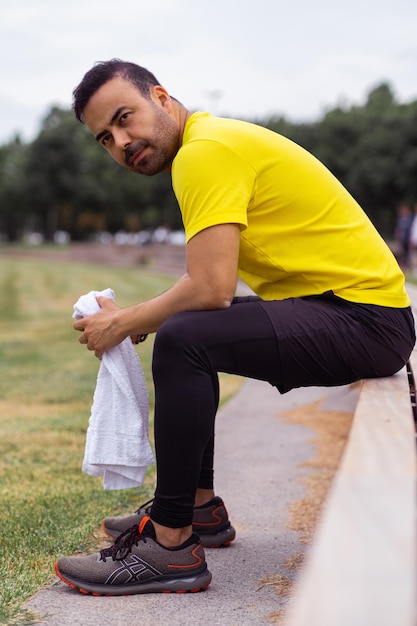 With towel in hand sporty man on a wooden bench in green city park taking a break