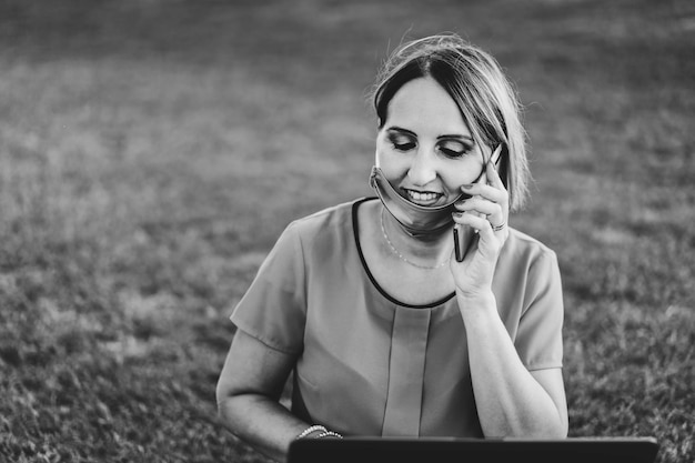 Woman 40 years old working outdoors with laptop during coronavirus outbreak Smiling entrepreneur sitting on lawn with personal computer while calling with cellphone Remote work concept into nature