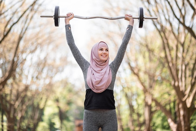 Woman in activewear and hijab holding barbell over head