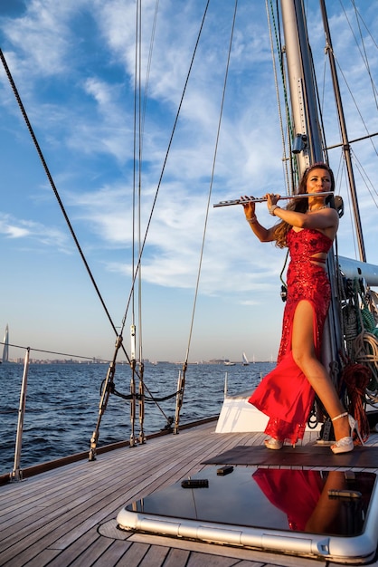 Woman actress in red stage costume with flute playing on yacht at sea and blue sky background Female in theatre clothes outdoors