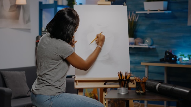 Woman of african american ethnicity drawing vase on white canvas and easel at studio. Black young artist working with art tools and pencil for modern masterpiece concept and fine art